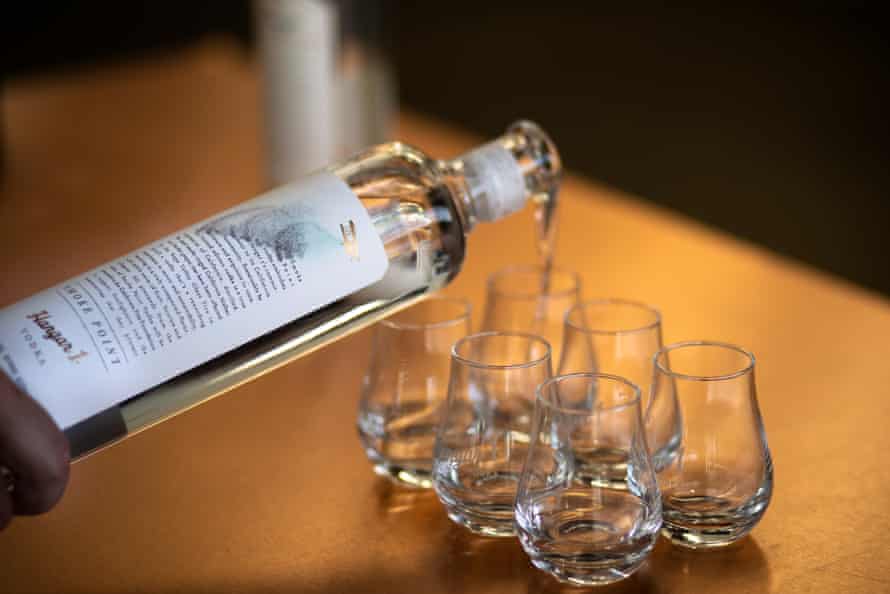 A bottle of Smoke Point vodka is being poured into six glasses lined up on a bar.