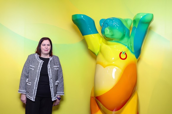 A woman in front of a yellow background. Next to her is the statue of the Berlin Bear with Fruit Logistica branding.