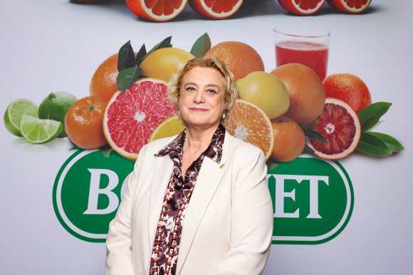 A woman stands in front of a large logo with lots of oranges and looks into the camera. 