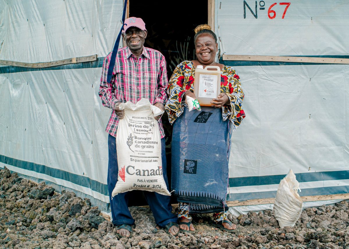 A smiling man and woman stand at the doorway of a tent shelter. The man holds a sack of what looks like grain and the woman holds a jug of cooking oil.