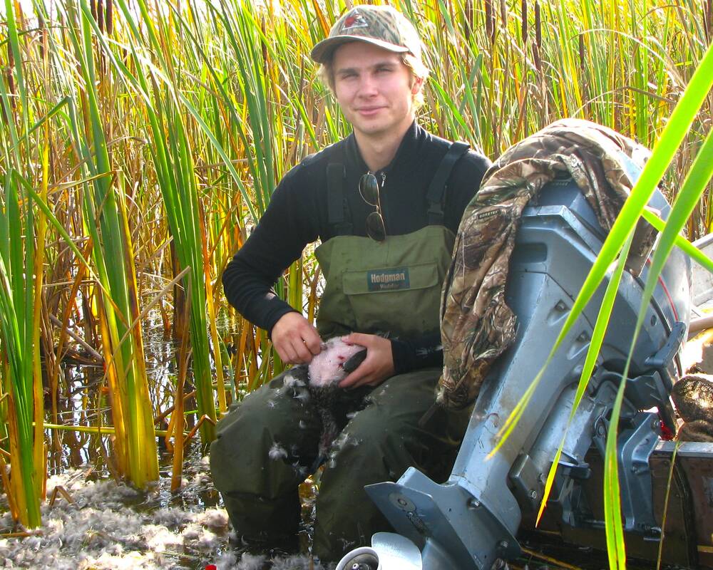 A young man sits on the edge of a boat on shore, next to its outboard motor, plucking a duck