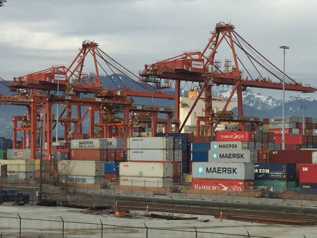 A shot of containers and the cranes that move them on the dock at the Port of Vancouver with the snow-capped mountains on the north shore behind them.