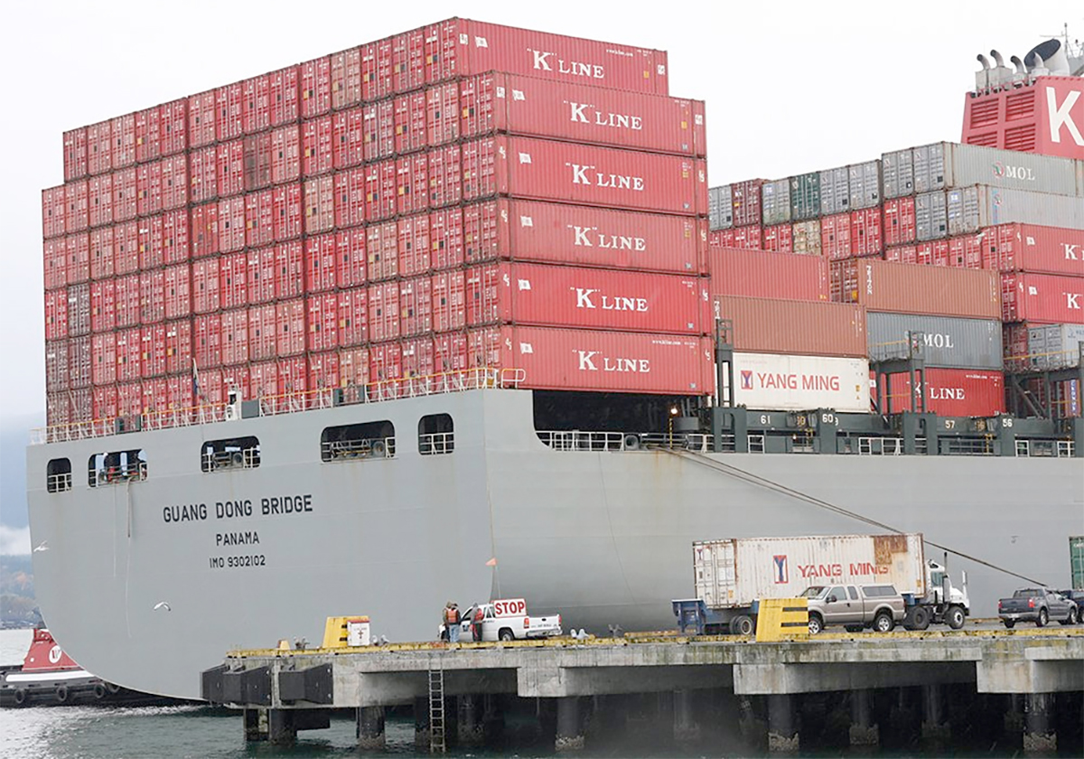 A massive grey ship at port in Vancouver has red shipping containers stacked six-high on its deck.