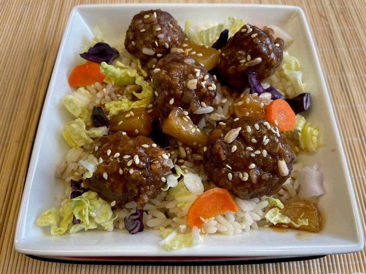 A plate with traditional sweet and sour meatballs on a bed of rice with some small bits of lettuce, pineapple, carrots and sesame seeds.