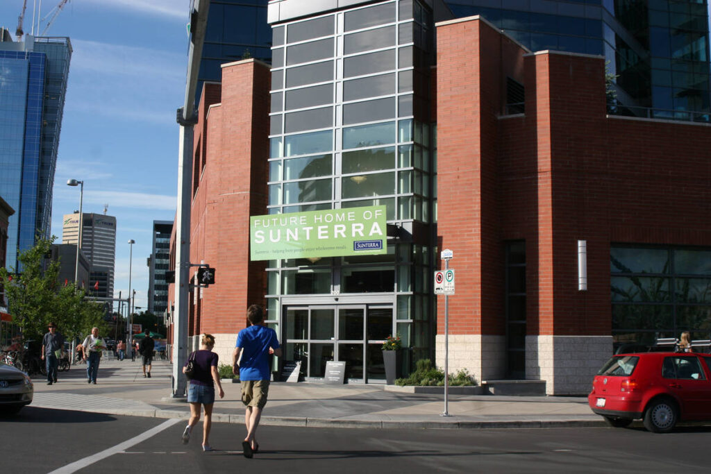 Two people cross a street walking toward a building on a corner with a sign on it that reads, 