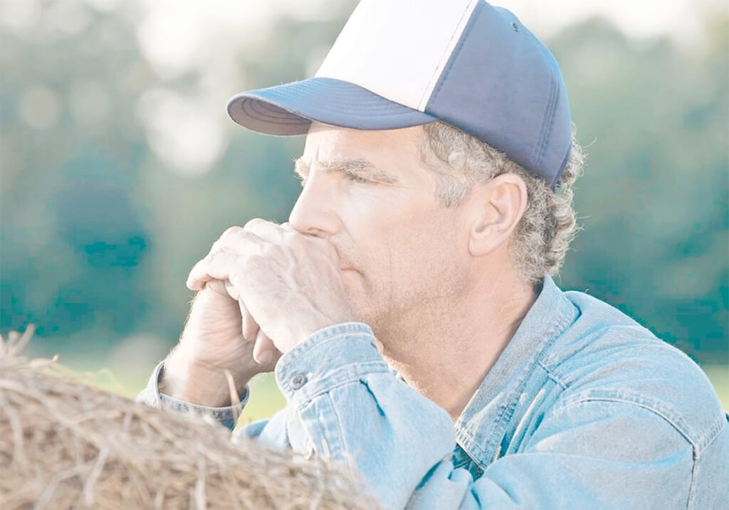 A stock photo of a farmer wearing a baseball cap, leaning on a large round bale, deep in thought.
