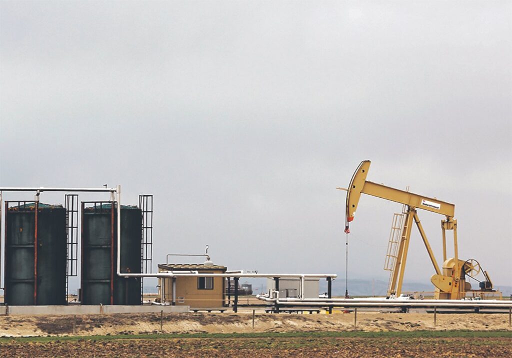 A crude oil pump jack stands in a field next to a couple of large black oil holding tanks.
