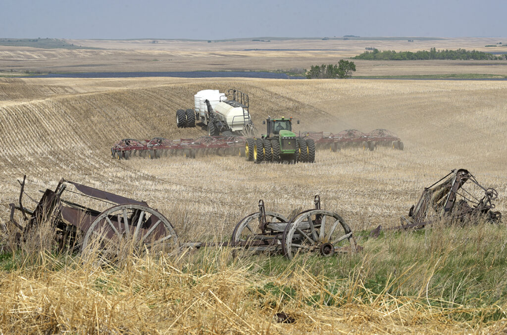 Old wooden farm wagons and wheels sit discarded in the foreground while a modern air seeder rig plants seed on a hillside in the background.