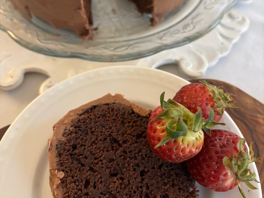 A piece of chocolate cake sits on a plate garnished with fresh, whole strawberries with the whole cake in the background.