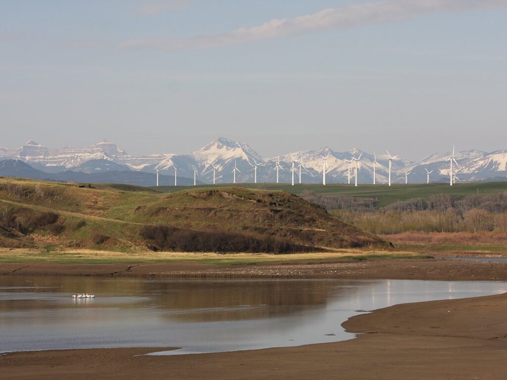 A scenic photo overlooking a small valley with some water and waterfowl in it, a ridge in the background with some wind turbines on it, and the snow-capped Rockies in the background.