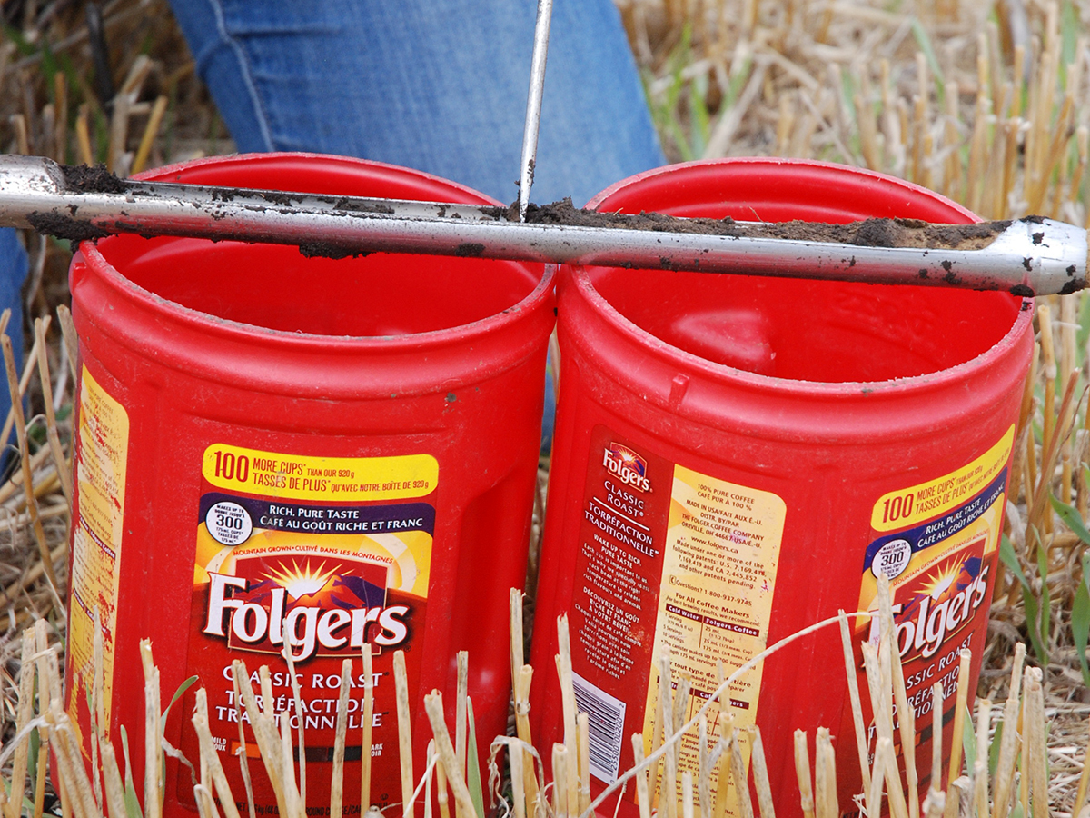 A soil probe rests on top of two red, plastic Folgers coffee containers.