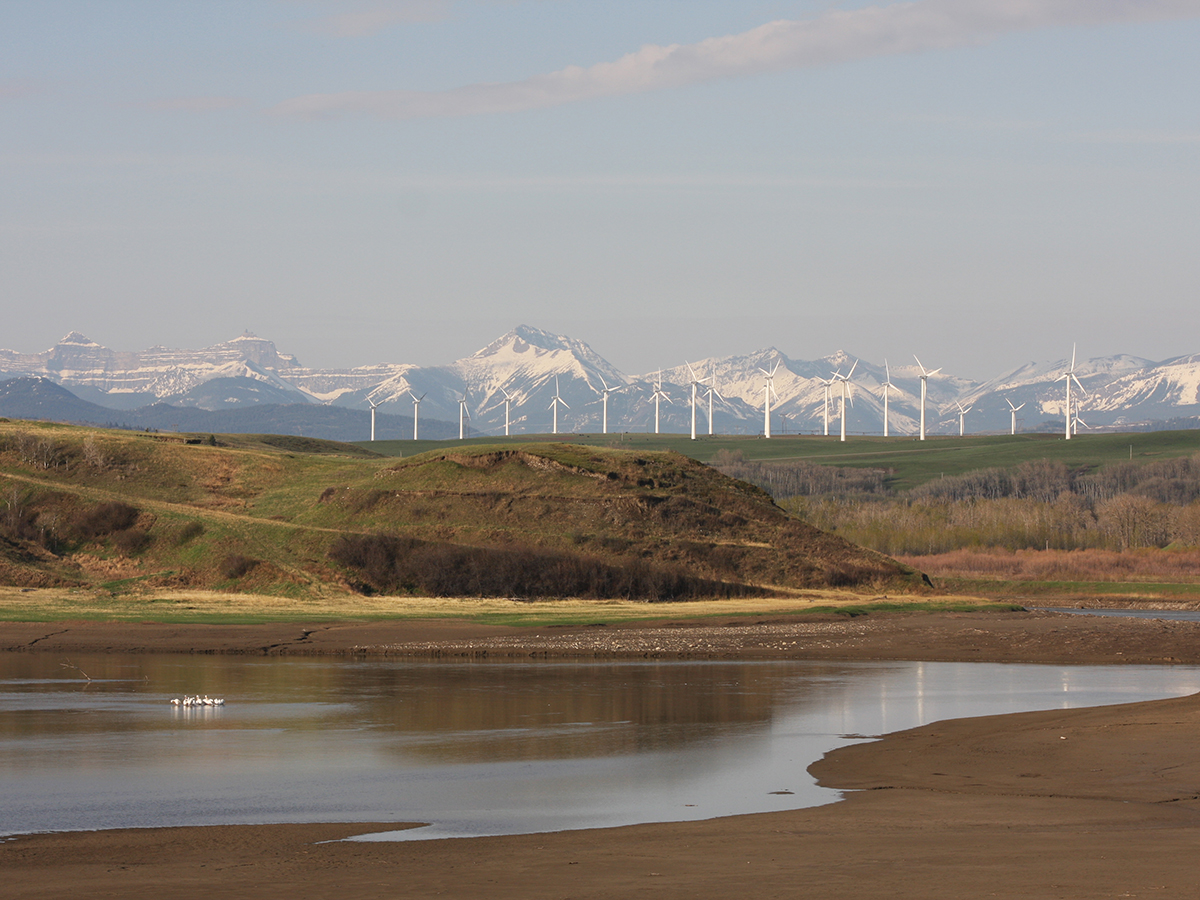 A scenic photo overlooking a small valley with some water and waterfowl in it, a ridge in the background with some wind turbines on it, and the snow-capped Rockies in the background.