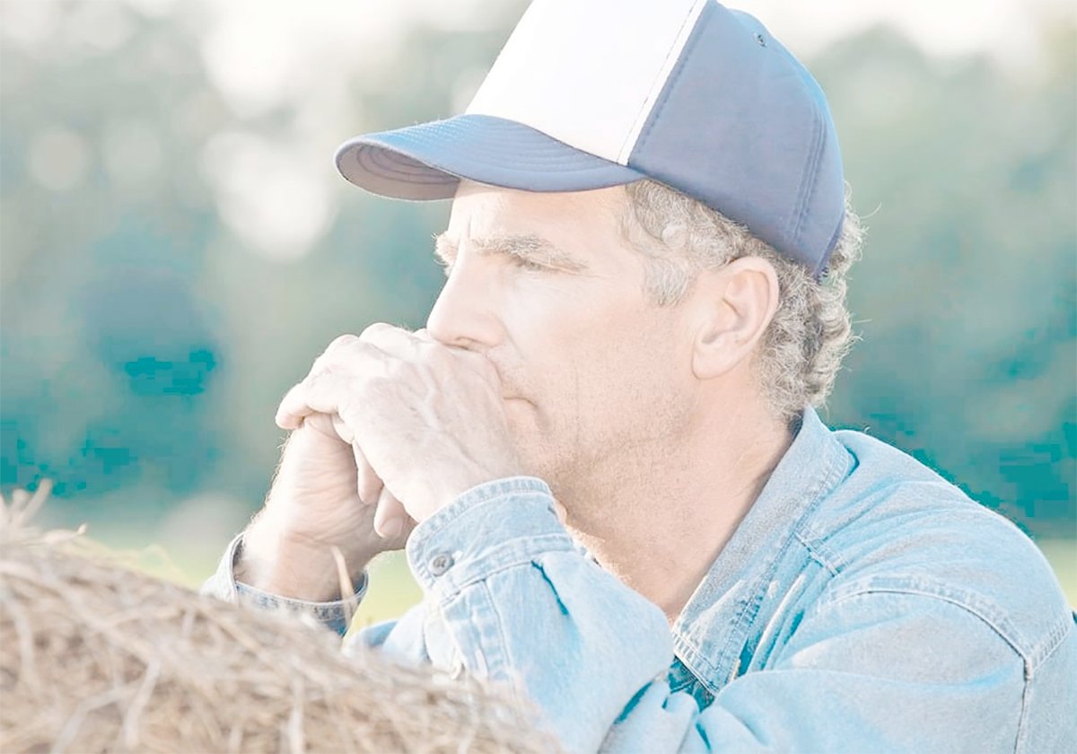 A stock photo of a farmer wearing a baseball cap, leaning on a large round bale, deep in thought.