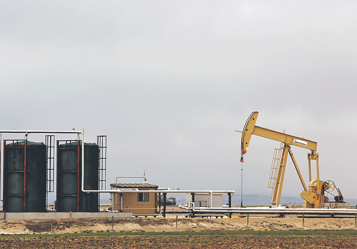 A crude oil pump jack stands in a field next to a couple of large black oil holding tanks.