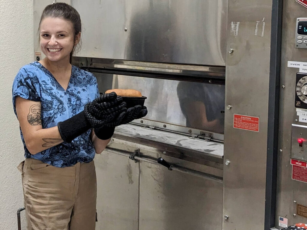A UC Davis researcher stands in front of an oven smiling and holding a loaf of bread made with a type of wheat that has had certain gluten proteins responsible for severe reactions in people with celiac disease deleted.