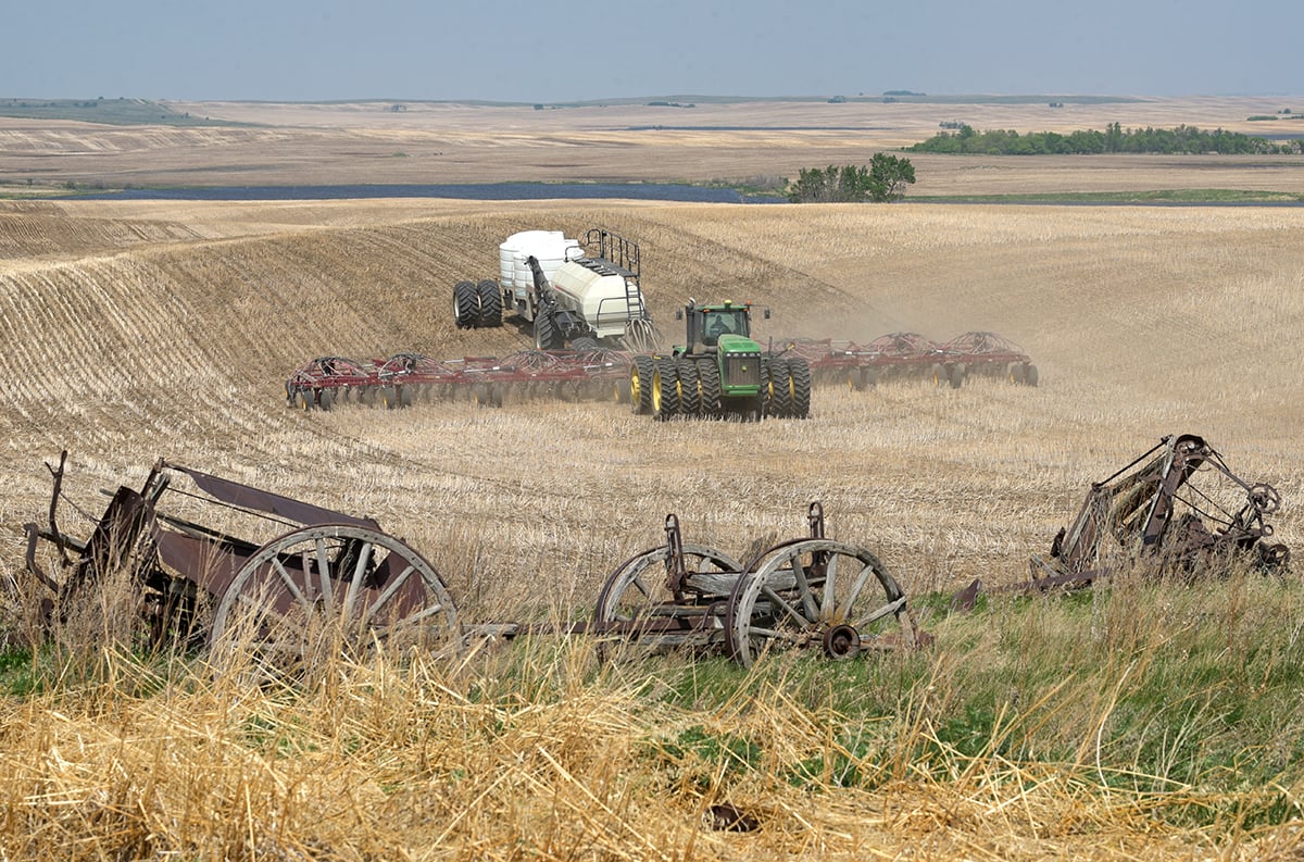 Old wooden farm wagons and wheels sit discarded in the foreground while a modern air seeder rig plants seed on a hillside in the background.