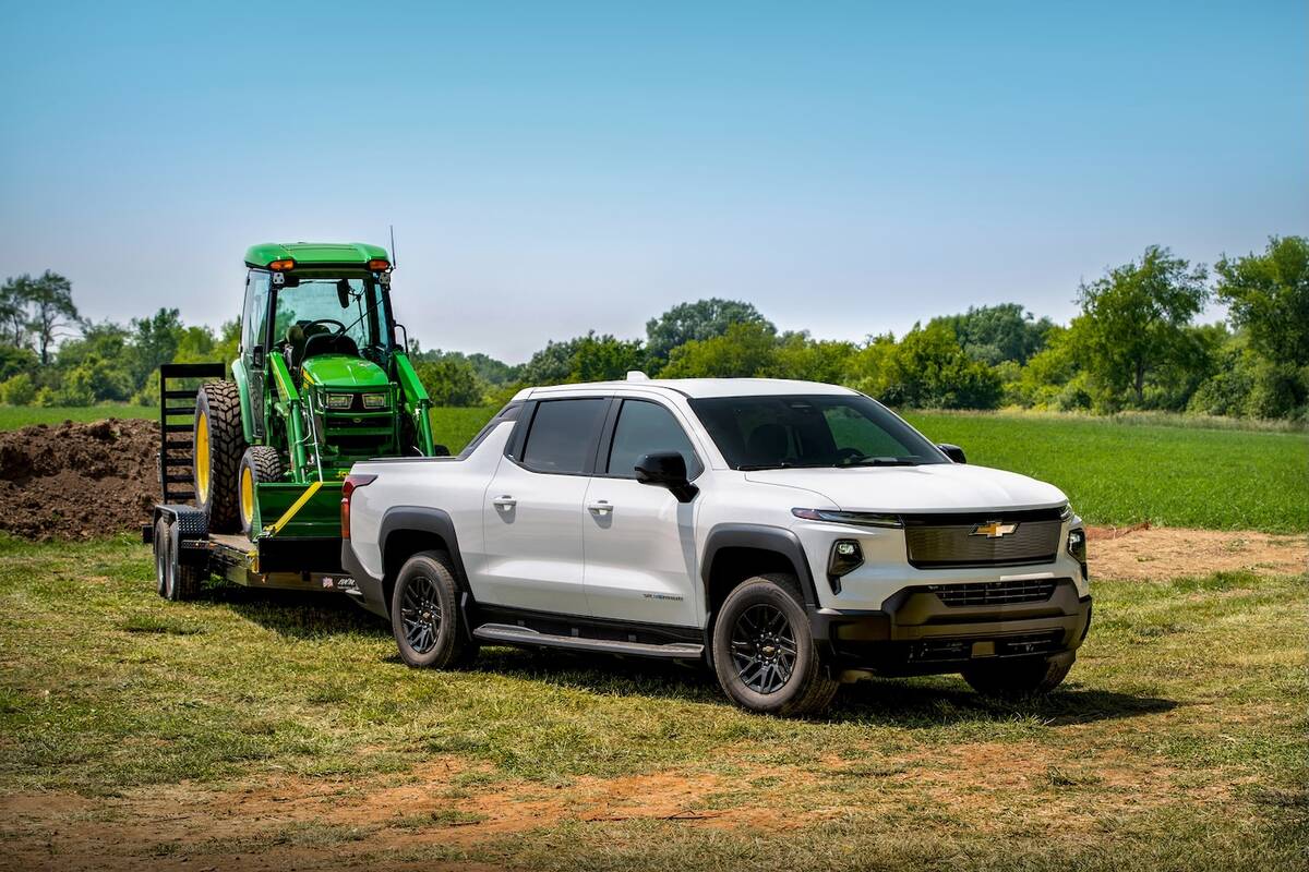 A white Silverado EV pulls a trailer with a John Deere utility tractor with a front end loader on it.