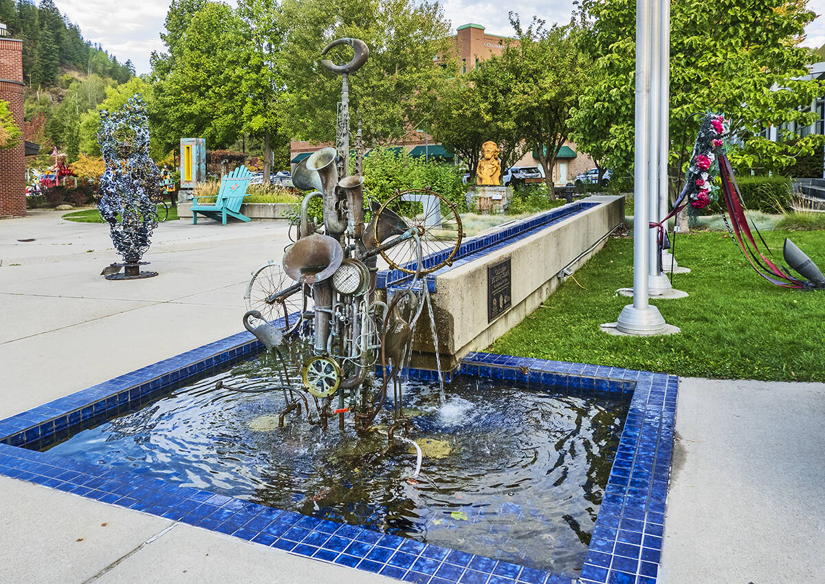 A unique sculpture made of old wind instruments like trumpets and tubas, along with bike wheels and a clock, serves as a fountain in the middle of a shallow pool in a public space.