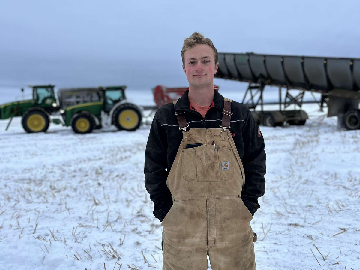 Mackenzie Fingerhut of Fingerhut Farms in Fairview, Alberta, poses in his coveralls for a photo with a couple tractors and snow-covered ground in the background.