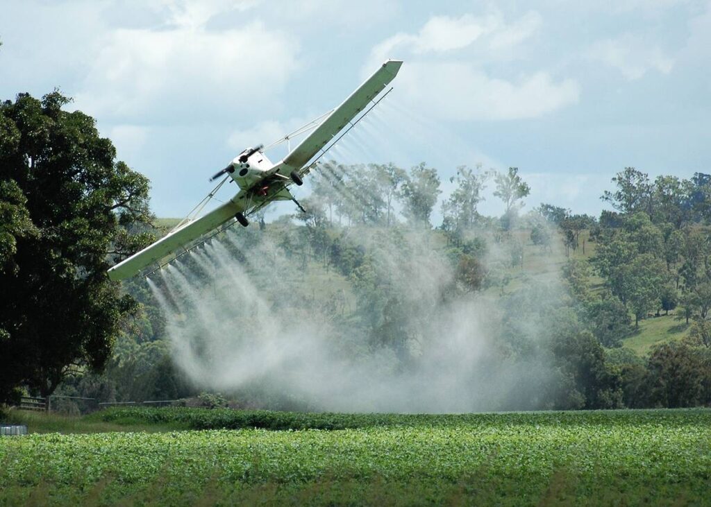 An aerial crop spraying plane flying low to the crop banks and climbs as it finishes a pass on a field surrounded by trees.