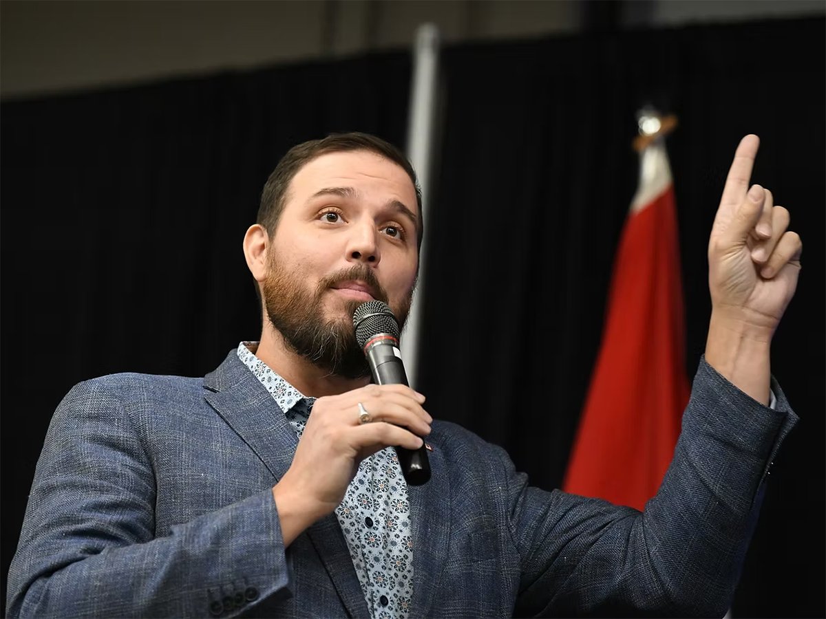 Damien Kurek speaks into a microphone while holding his left index finger up pointing to the sky.