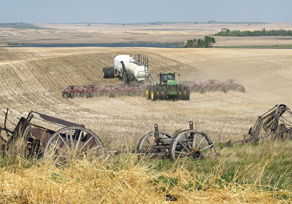 Photo looking down from the top of a hill with a modern air seeder rig approaching in the distance and some discarded old wooden equipment with wagon wheels in the foreground. with