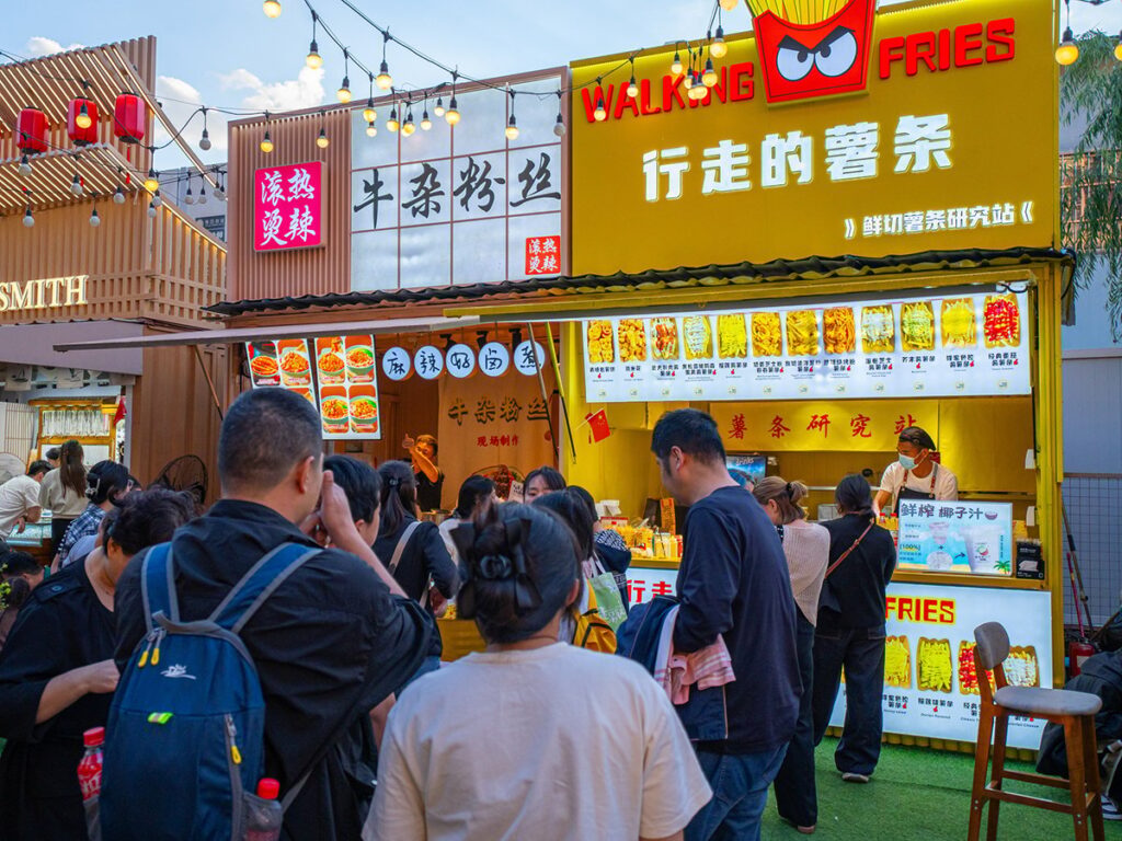 A crowd of people in a Chinese market with food vendor booths in the background, one called, 