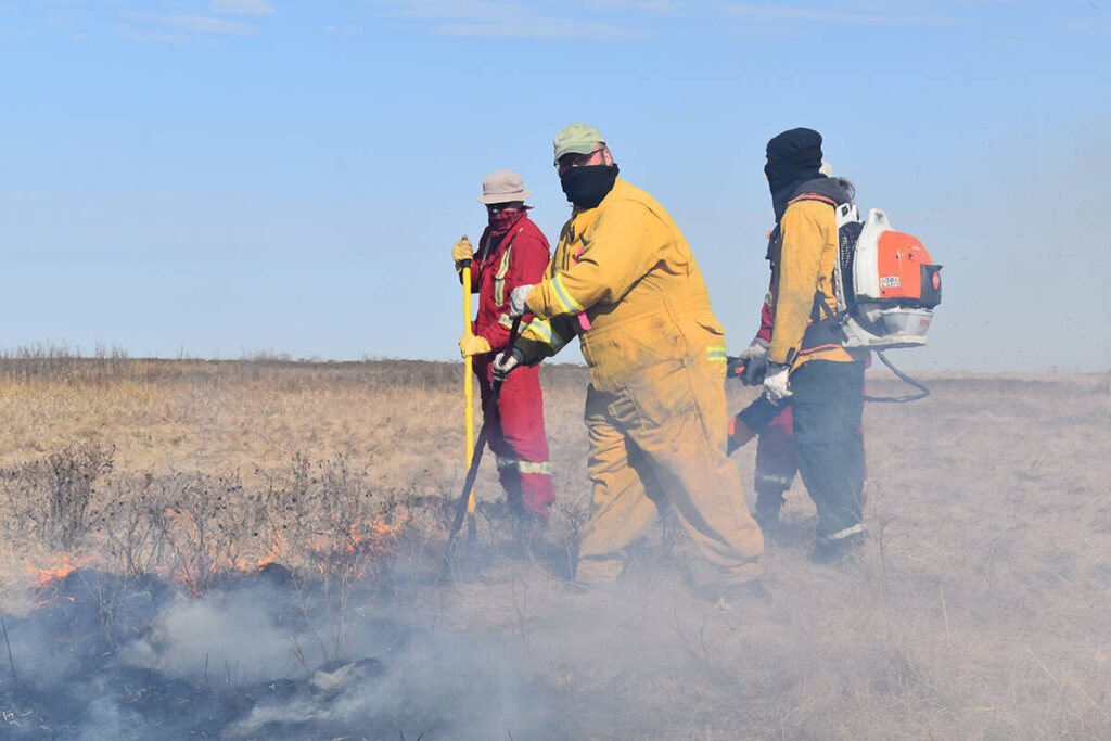 Four people clad in red and yellow fire-resistant coveralls, wearing face-coverings and holding hand tools while one wears a backpack leaf blower, scrape at the charred ground cover in a pasture with smoke rising around them.