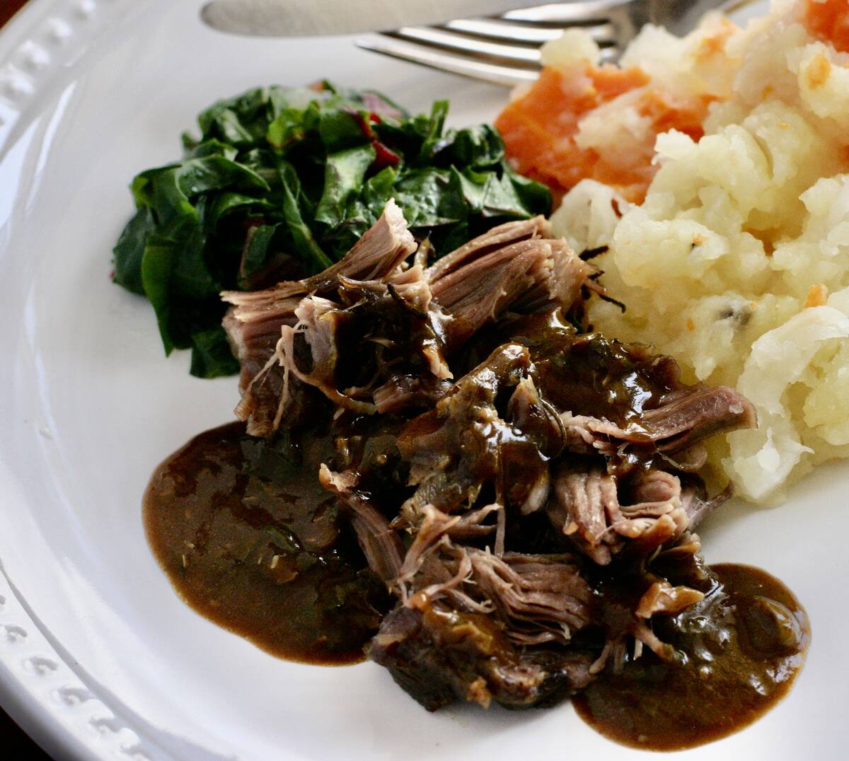 Shredded roasted lamb shoulder with thick, dark gravy on top sits on a plate next to some mashed potatoes.