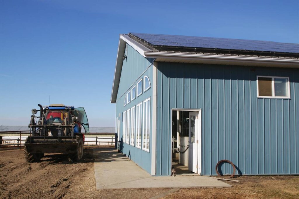 A front-wheel-assist tractor with a front-end loader with grapple on it is parked outside a newer farm shop building with lots of windows in it and solar panels on its roof.