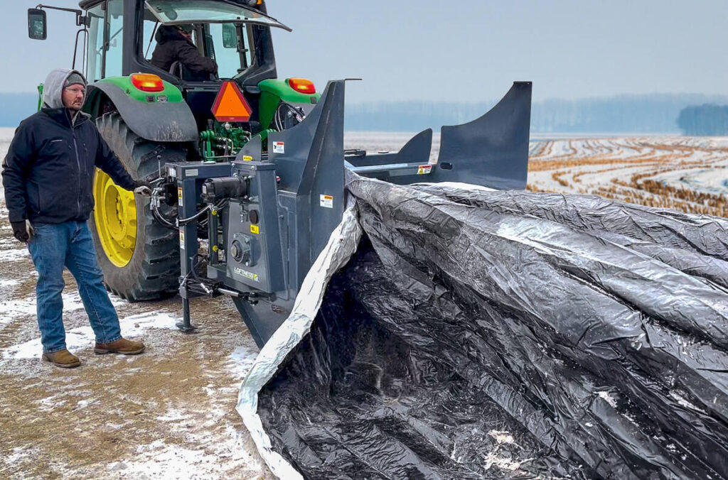 A farmer stands beside a tractor-mounted grain bag roller.