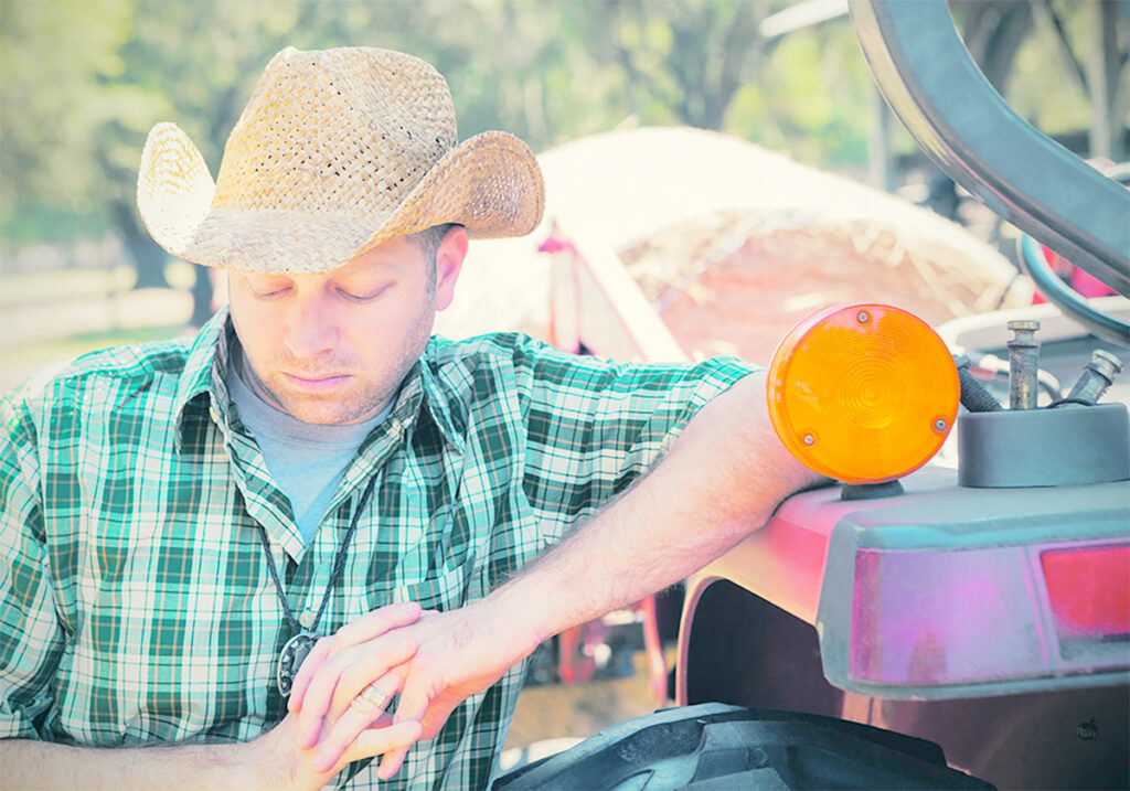 A stock photo of a young farmer in a cowboy hat leaning against his tractor and staring at the ground.
