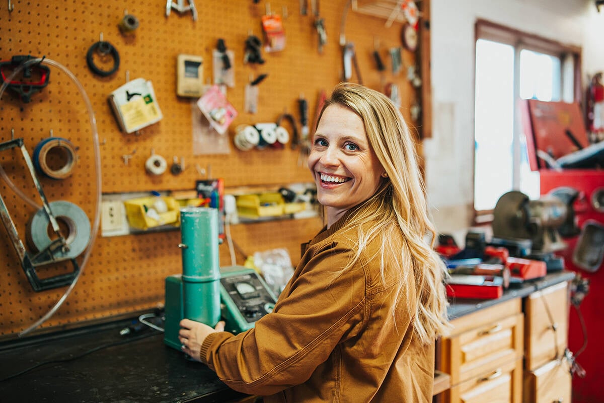 Lesley Kelley smiles for a photo while working with her grain tester in her shop, tools hang on the peg board wall in the background.