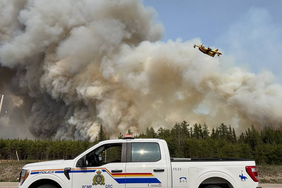 An RCMP truck is in the foreground while a huge column of smoke from wildfires rises in the background, and a water bomber aircraft passes over top of it.