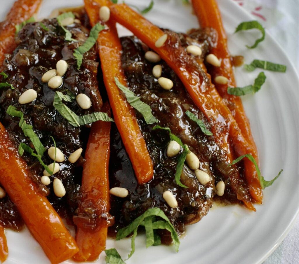 A glazed lamb steak sits on a plate with slivers of carrot, pine nuts and fresh basil leaves on top.