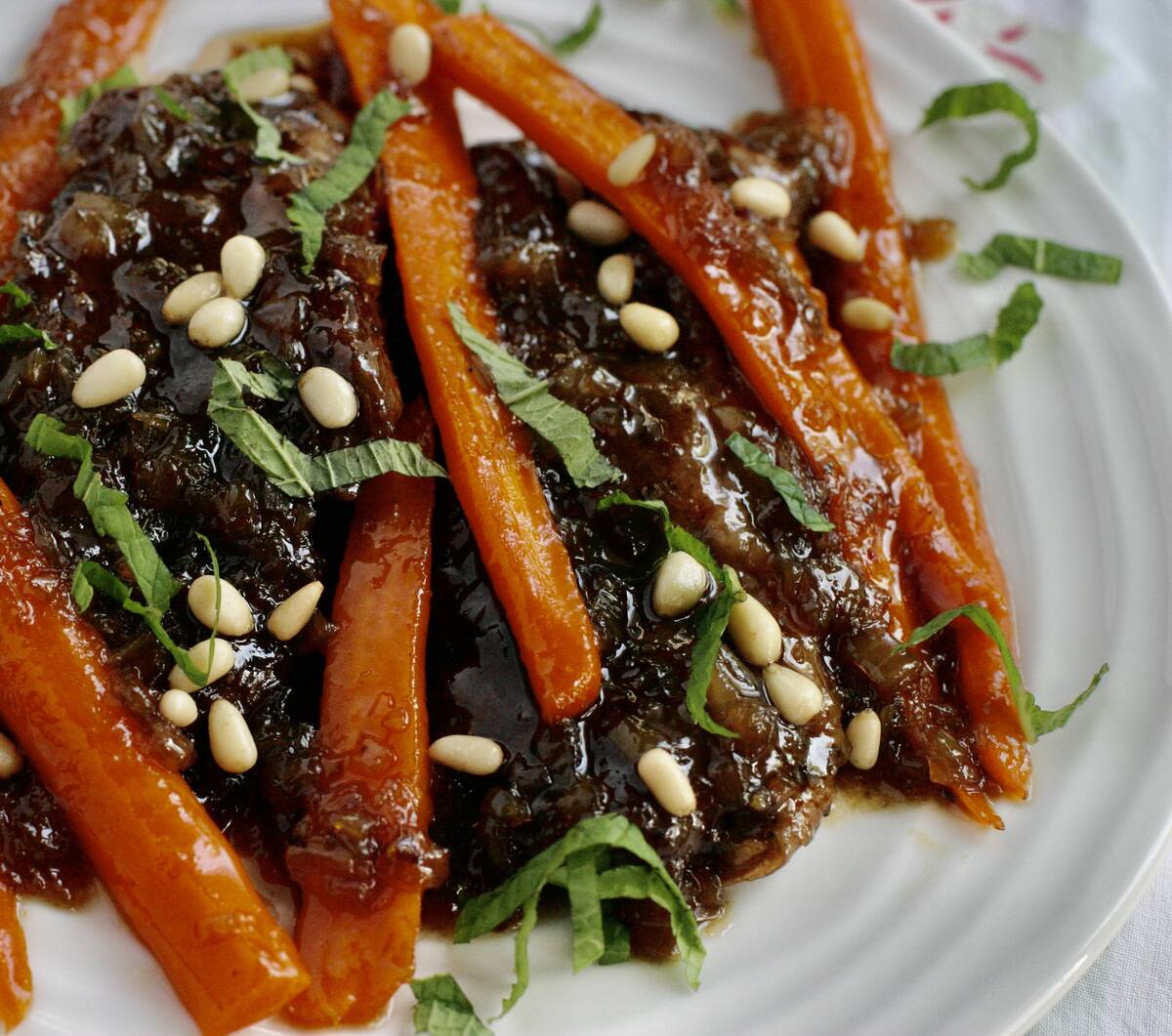 A glazed lamb steak sits on a plate with slivers of carrot, pine nuts and fresh basil leaves on top.