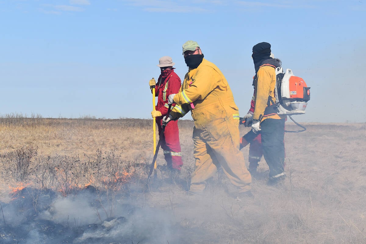 Four people clad in red and yellow fire-resistant coveralls, wearing face-coverings and holding hand tools while one wears a backpack leaf blower, scrape at the charred ground cover in a pasture with smoke rising around them.
