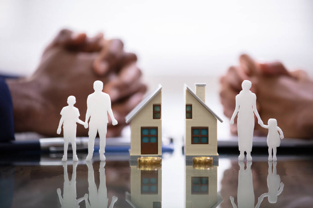 A stock image of a model of a house, split in half, sitting atop a table with the silhouettes of the parents, each holding the hand of a child, are on opposite sides of the divided home.