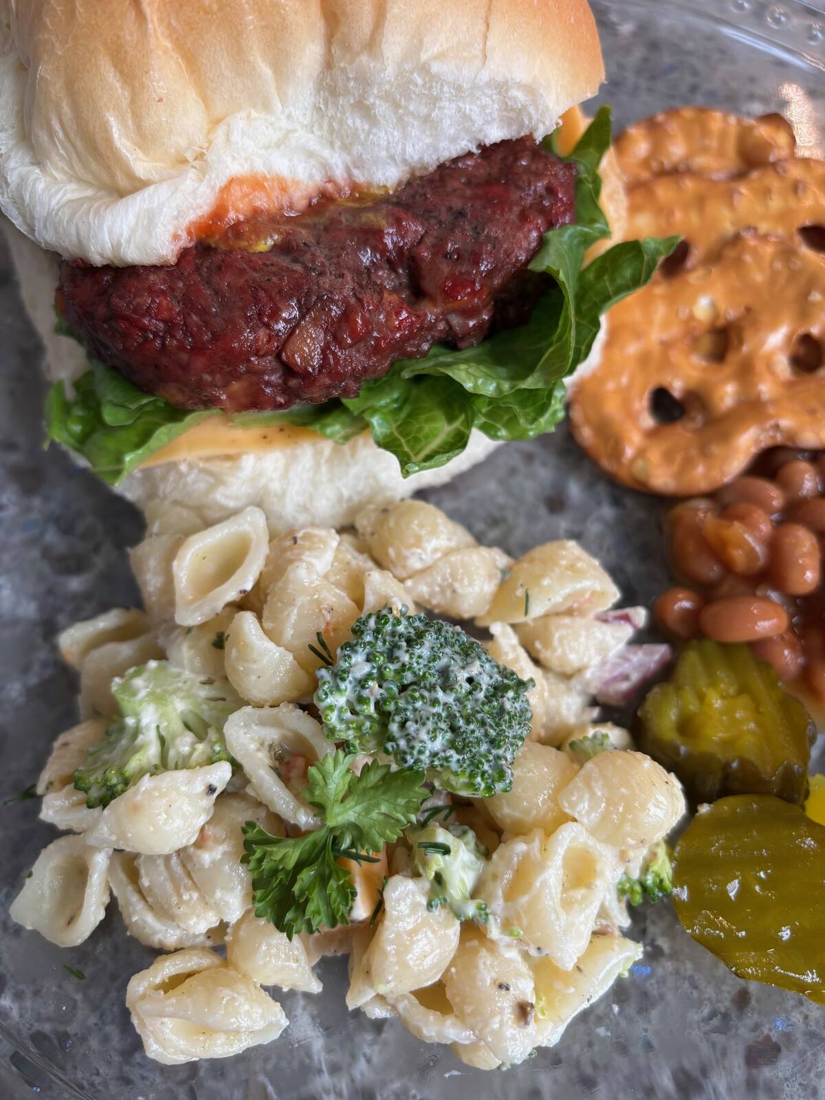Seashell pasta in white sauce with a sprig of broccoli on top on a plate next to a burger in a bun, some pretzel chips and a few sliced pickles.