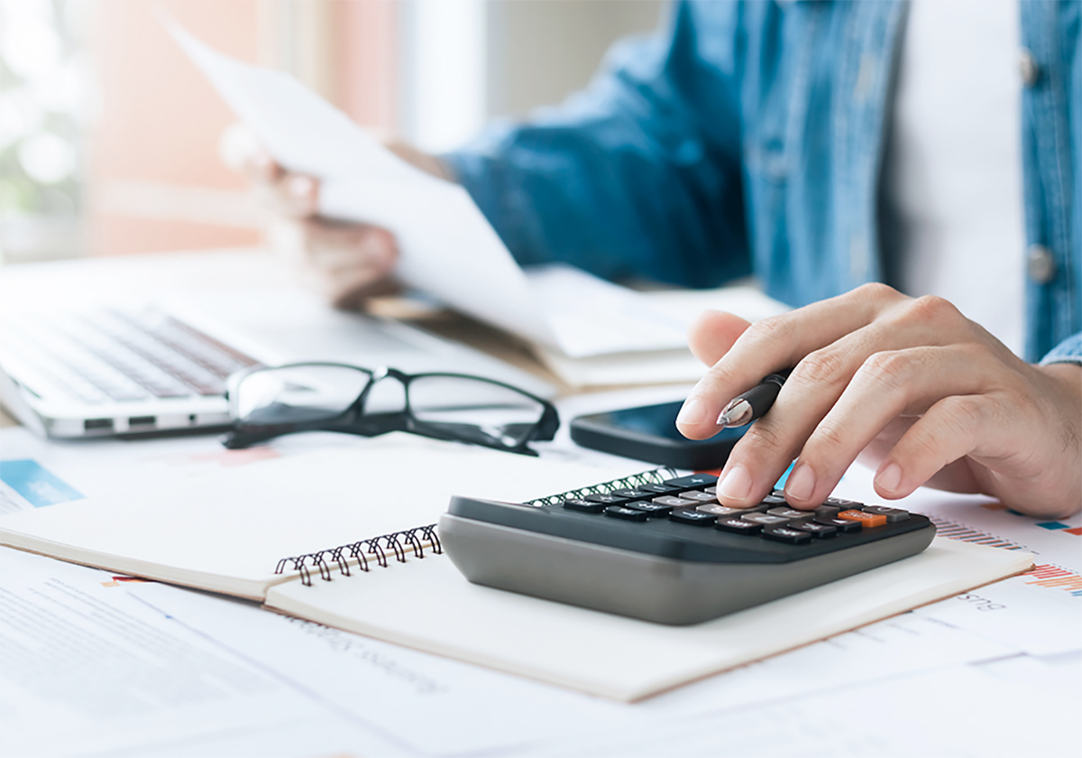 A man's left hand works on a calculator while his right holds a paper. A laptop computer and reading glasses are also on the desk.