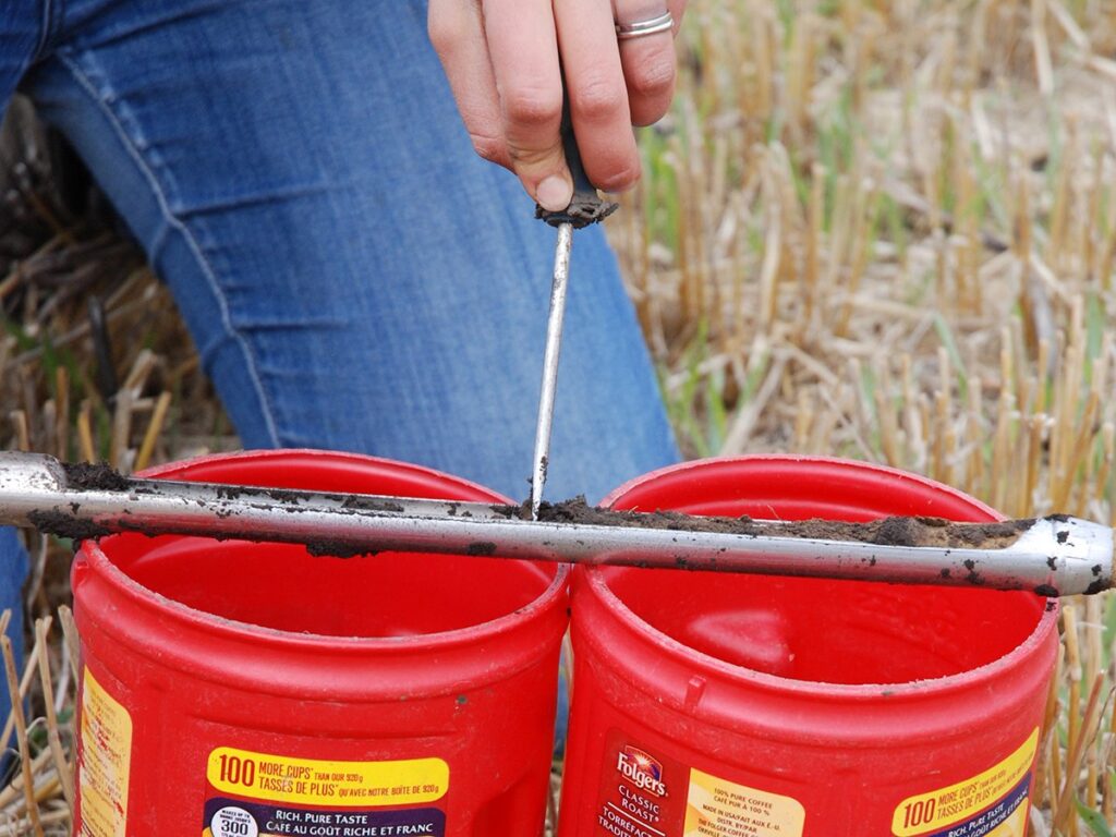 A hand uses a tool to scrape soil from a probe into two red, plastic coffee containers in a field.