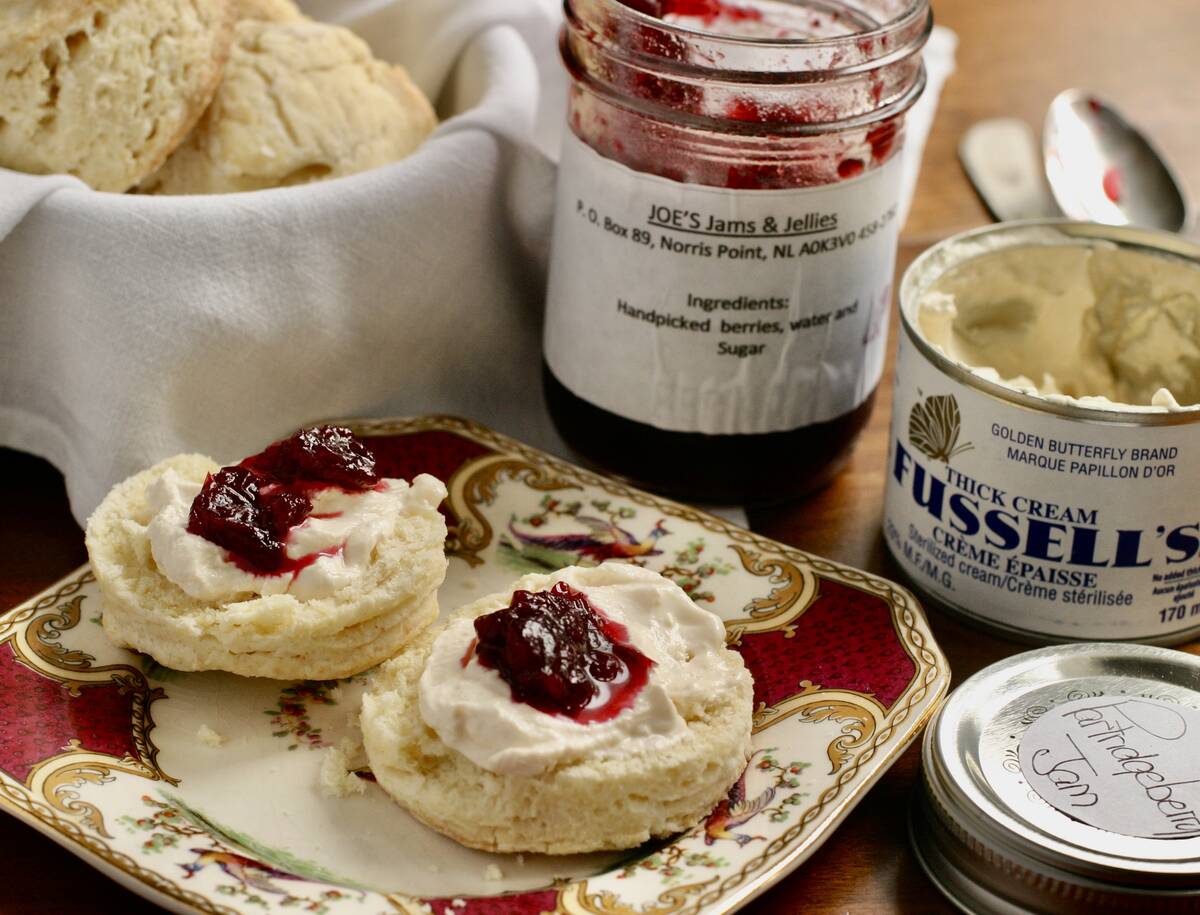 Two scones topped with thick cream and jam sit on a decorative plate with more behind them in a basket.