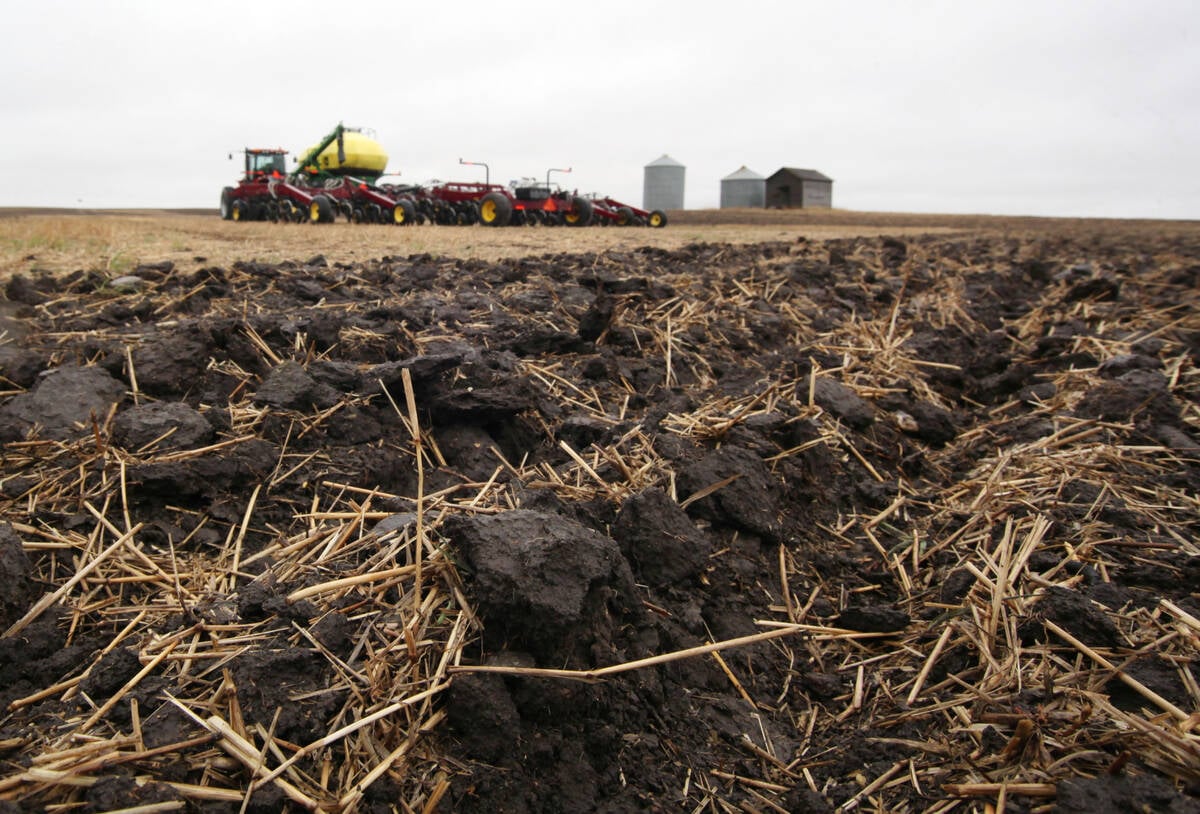 A low angle photo of some disturbed soil with some litter on top of it and an air seeder parked in the background on a cloudy day.