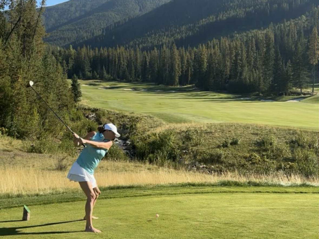 A woman in bare feet tees off on a scenic golf course in the mountains.