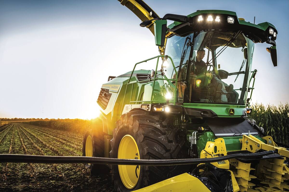 A John Deere forage harvester sits with a farmer at its controls while the sun sets immediately behind the machine.