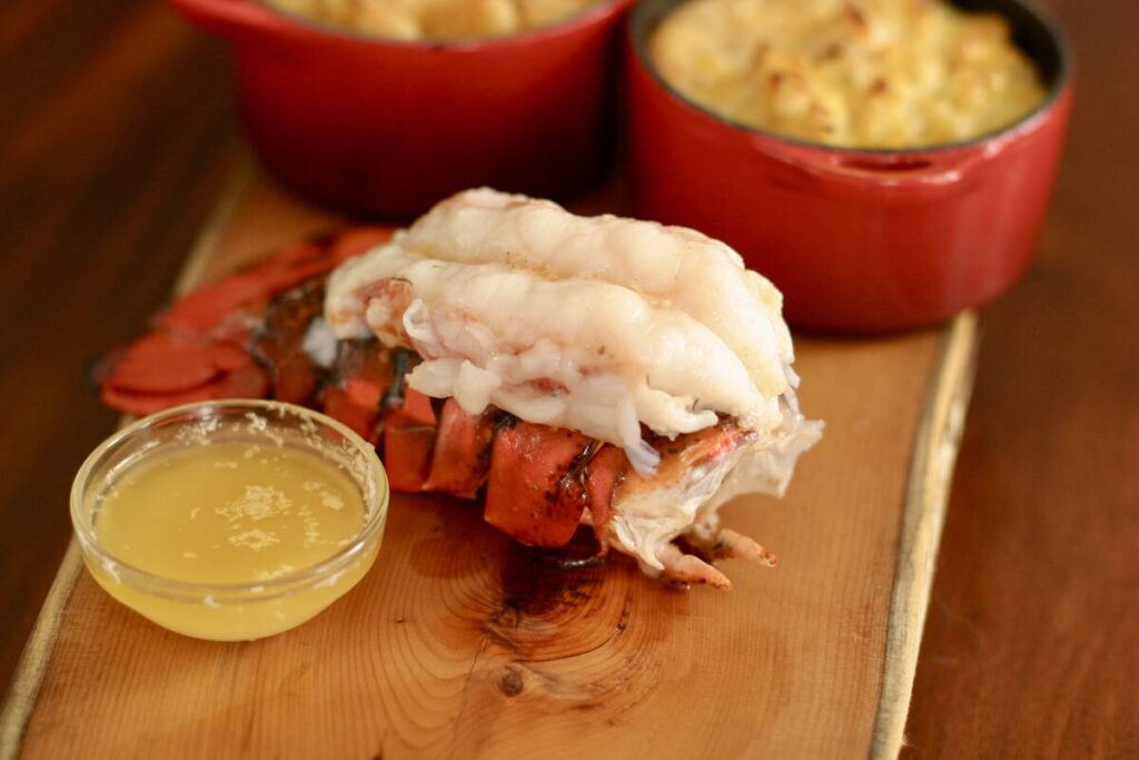 A freshly-cooked lobster tail sits on a wooden table with a small bowl of melted butter beside it.