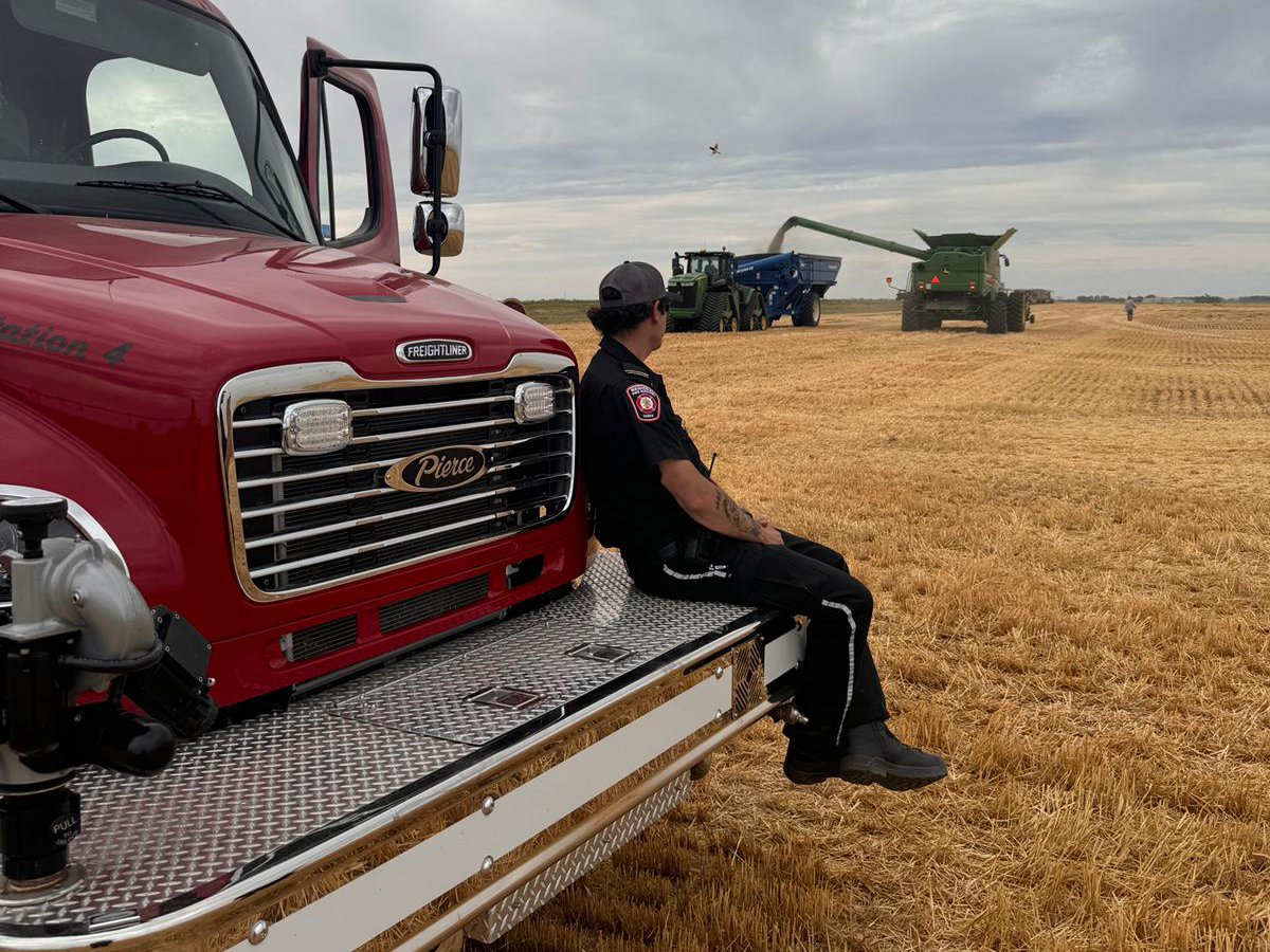 A volunteer firefighter in station wear sits on the front bumper of a pumper truck parked in a field with a combine augering crop into the box of a grain truck in the background.
