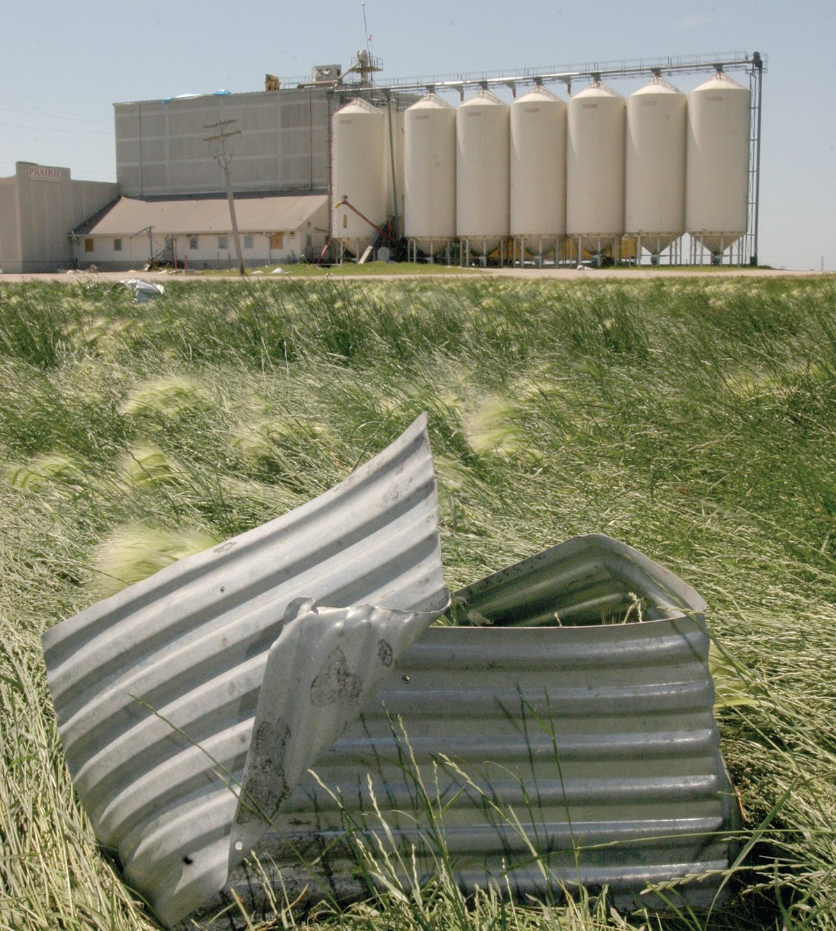 A piece of twisted corrugated metal sits on the grass with an inland grain terminal in the background.