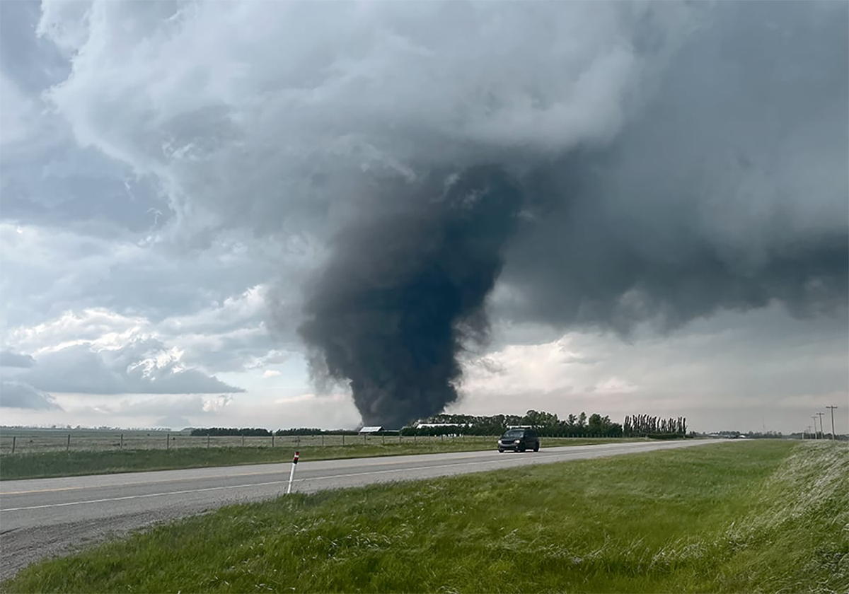 A vehicle drives by on a highway in the foreground while a dark and massive tornado spins in the background.