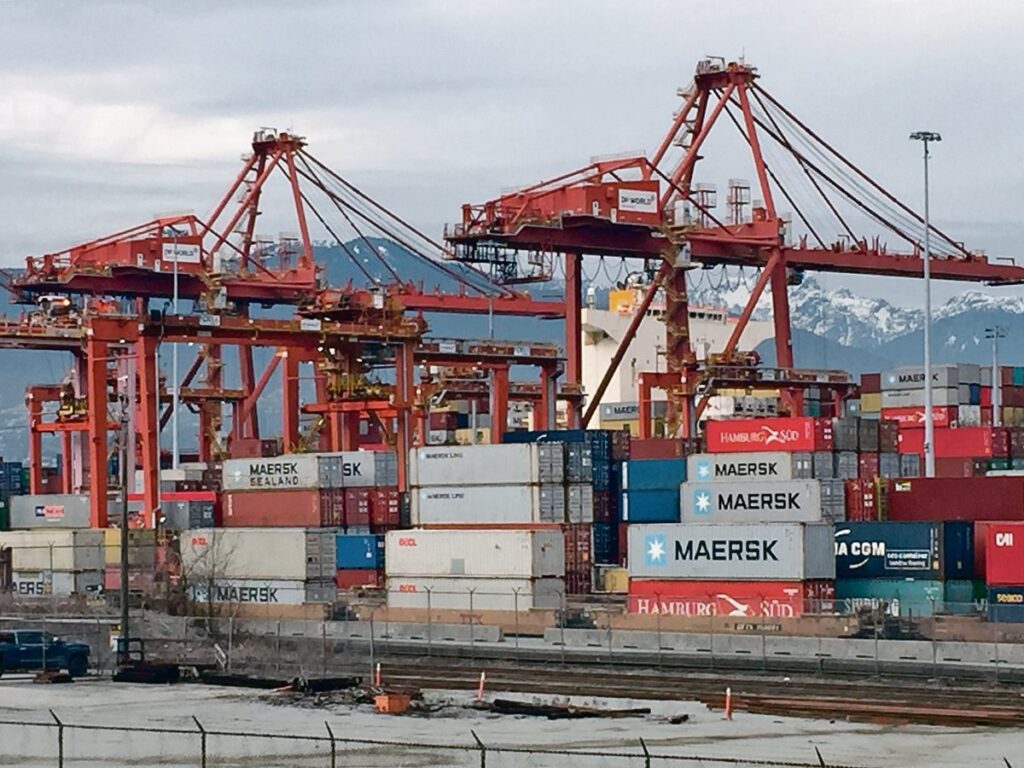 Stacks of shipping containers sit dockside beneath the massive cranes that hoist them aboard ships in the Port of Vancouver with the mountainous North Shore visible in the background.